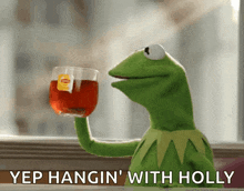 kermit the frog is holding up a glass of lipton tea and says yep hangin ' with holly