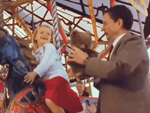 a man holds a teddy bear while a little girl rides a carousel