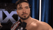a shirtless man with a beard holds a microphone in front of a large x