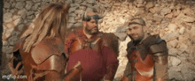 a group of people in armor are standing next to each other in front of a stone wall talking to each other .