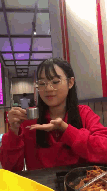 a girl wearing glasses is sitting at a table holding a small container