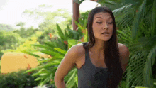 a woman in a tank top is standing in front of a lush green garden .
