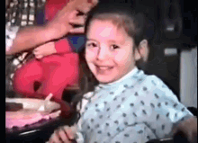 a little girl is sitting at a table with a cake in the background and smiling .