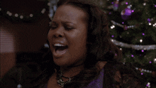 a woman wearing a purple dress and a necklace is smiling in front of a christmas tree