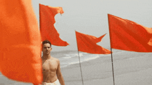 a shirtless man stands in front of three red flags on the beach