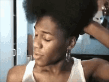 a woman in a white tank top adjusts her hair in front of a mirror .