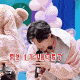 a man in a pink jacket is holding a microphone in front of a teddy bear and balloons
