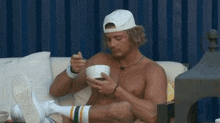 a shirtless man is sitting on a couch eating from a bowl with a fork .