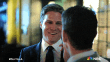 a man in a suit and tie smiles while talking to another man with #suitsla written on the bottom