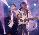 a man singing into a microphone while another man plays a guitar in front of a drum set that says rock on it