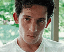 a man with curly hair is wearing a white t-shirt and looking at the camera with a serious look on his face .