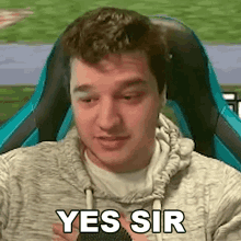 a man in a grey hoodie is sitting in a blue chair and making a yes sir gesture .