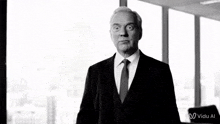 a man in a suit and tie is making a funny face in front of a window with a vidu al logo in the corner
