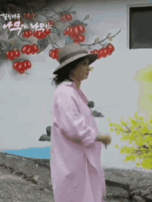 a woman in a pink coat and hat is standing in front of a wall with a painting of a tree on it .