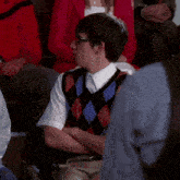 a young man wearing glasses and a plaid vest sits in a crowd of people