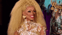 a drag queen with very long blonde hair is wearing a white and gold dress .