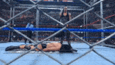 a wrestler is laying on the ground in a cage while a referee stands behind him .