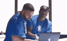 two men in blue shirts are looking at a laptop computer .