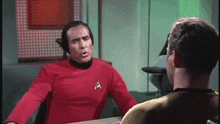 a man in a red shirt with a star trek logo on his chest is talking to another man in a yellow shirt .