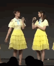 two women in yellow dresses are standing next to each other on a stage singing into microphones .