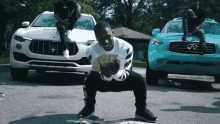 a man is squatting down holding a fan of money in front of a maserati and an infiniti .