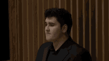 a young man in a black suit is sitting in front of a wooden wall