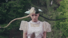 a woman in a white dress with two braids in her hair is standing in the woods .