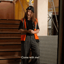 a man with long hair wearing a hard hat and an orange vest says come with me