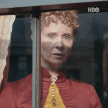 a woman in a red shirt and yellow tie looks out a window with hbo written on the bottom