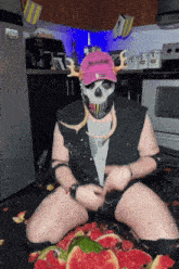 a man wearing a skull mask and a pink hat with x on it sits on the floor surrounded by watermelon slices