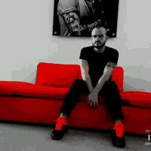 a man with a tattoo on his arm is sitting on a red couch in front of a picture of a man