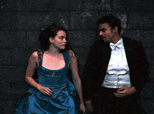 a man in a tuxedo and a woman in a blue dress lean against a wall