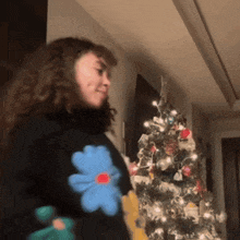 a woman standing in front of a christmas tree wearing a black sweater with flowers on it