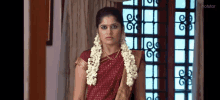 a woman in a red saree is standing in front of a window with a garland of flowers around her neck .