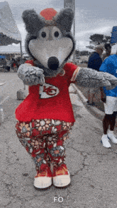 a mascot dressed as a wolf wearing a red shirt and floral pants is standing on the street .