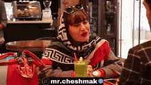a woman in a scarf is sitting at a table drinking a green drink .