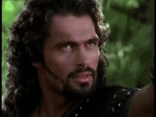 a man with long hair and a beard is looking at the camera in a forest .
