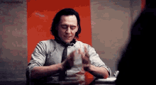 a man in a suit and tie is sitting at a table with his hands folded in front of him .