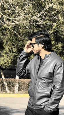 a man wearing a leather jacket and sunglasses stands in front of a tree