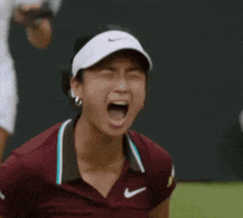 a woman wearing a nike visor and a maroon shirt is screaming on a tennis court