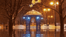 a gazebo in the middle of a snowy park with a reflection of it in the water