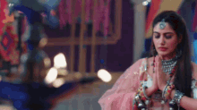 a woman in a pink dress and jewelry is praying with her hands folded .