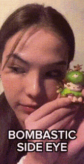 a woman is holding a green figurine with the words bombastic side eye on it