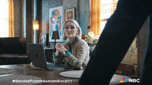 a woman sits at a table with a laptop and a cup of coffee and a nbc logo in the corner