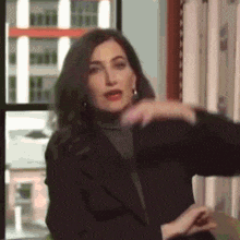 a woman in a black jacket is standing in front of a window and making a funny face