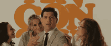 a man in a suit and tie is surrounded by three women in front of an orange sign that says con