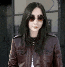 a woman wearing a leather jacket and round sunglasses is standing in front of a window .