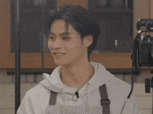 a young man wearing an apron and a white hoodie is smiling in a kitchen .