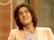 a man with long hair is wearing a white suit and striped shirt .