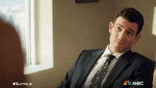 a man in a suit and tie is sitting in front of a window with #suitsla written on the bottom right